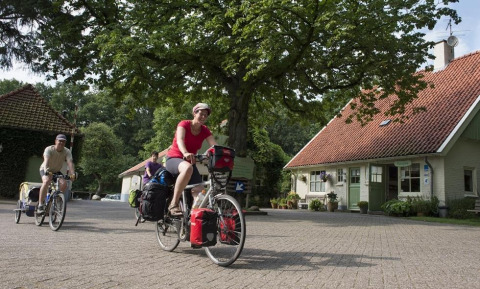 Ciclisti pedalano nel villaggio turistico Huttopia De Roos a Overijssel, Paesi Bassi, tra alberi e case.