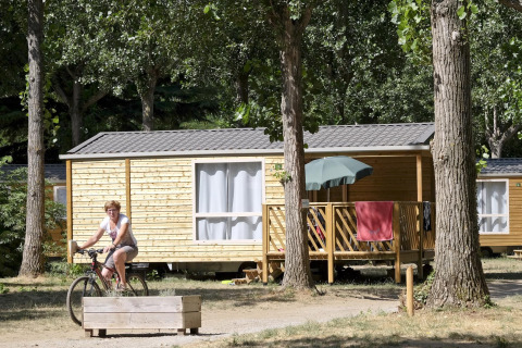 Kvinde cykler foran en Mobile home Cottage i skov, terrasse med parasol og håndklæder i solen.