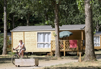 Kvinde cykler foran en Mobile home Cottage i skov, terrasse med parasol og håndklæder i solen.