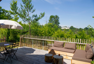 Terraza de madera con asientos y sombrilla, vista al bosque en Village Huttopia Lanmary, Nouvelle-Aquitaine, Francia.