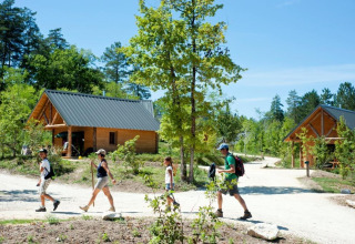Family hiking on a sunny trail with wooden cabins at Village Huttopia Lanmary holiday park, Nouvelle-Aquitaine, France.