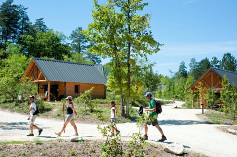 Family hiking on a sunny trail with wooden cabins at Village Huttopia Lanmary holiday park, Nouvelle-Aquitaine, France.