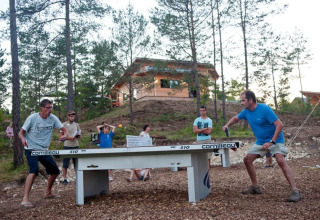Feriegæster spiller bordtennis udenfor blandt træer ved Village Huttopia Lanmary i Nouvelle-Aquitaine, Frankrig.