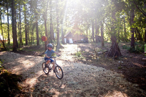 Niño con casco monta en bicicleta por un sendero boscoso cerca de cabañas en Village Huttopia Lanmary, Francia.