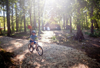 Barn cykler med hjelm på en skovsti i sollys nær hytter i Village Huttopia Lanmary, Frankrig.