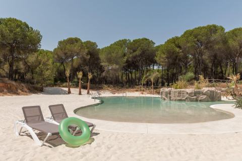 Swimming pool with sandy beach, lounge chairs, and trees in the background at Huttopia Parque de Doñana, Andalusia.