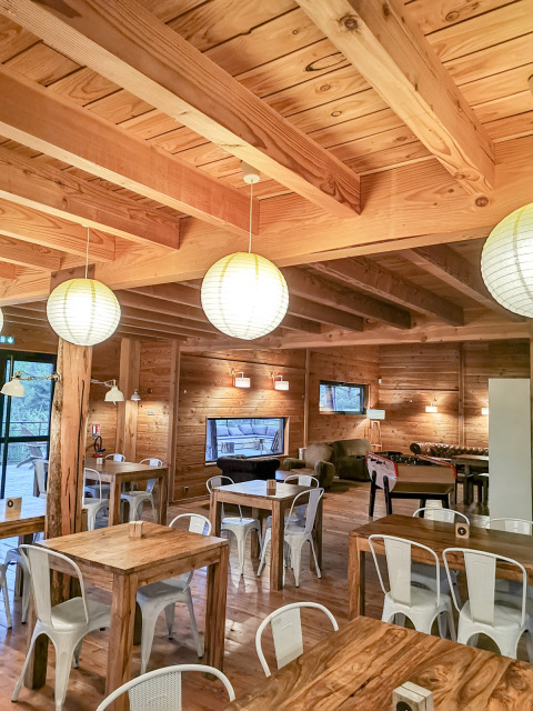 Area comune accogliente con tavoli in legno e sedie bianche al Village Huttopia Sud-Ardèche, Auvergne-Rhône-Alpes, Francia.