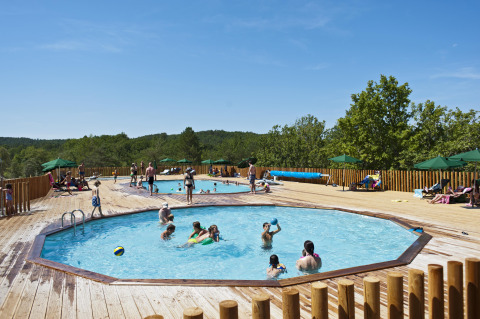Außenpools mit Gästen, Holzterrasse und grünen Sonnenschirmen im Village Huttopia Sud-Ardèche, Frankreich.