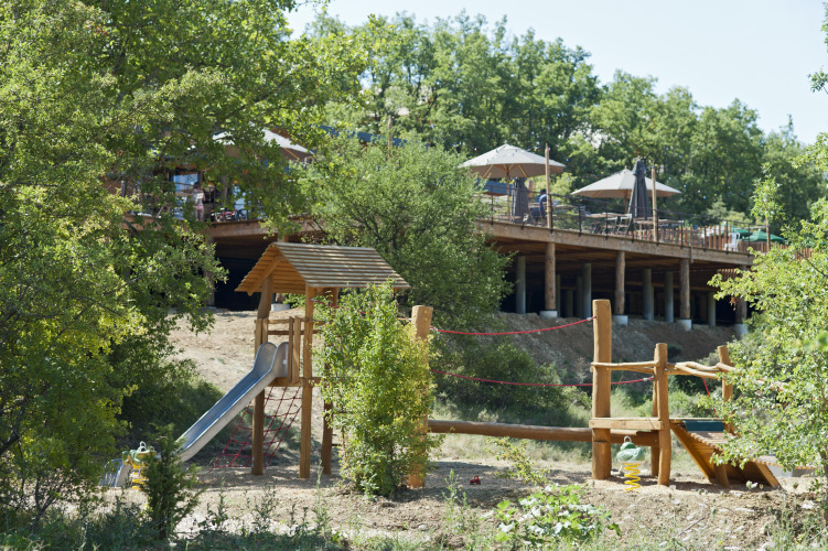 Aire de jeux extérieure et terrasse en bois avec parasols dans un parc de vacances en Ardèche, France.