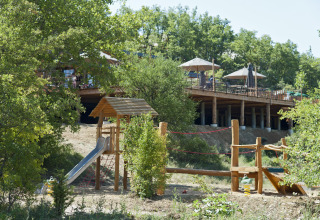 Spielplatz im Freien und Holzterrasse mit Sonnenschirmen in einer Ferienanlage in Auvergne-Rhône-Alpes.