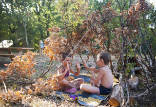 Zwei Kinder spielen gemeinsam in einer aus Ästen gebauten Hütte im Freien im Village Huttopia Sud-Ardèche.