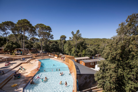 Piscina al aire libre con personas disfrutando junto a terrazas y cabañas de madera en Huttopia La Forêt de Janas.