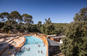 Udendørs swimmingpool omgivet af træer og træhytter på ferieparken Huttopia La Forêt de Janas i Provence, Frankrig.