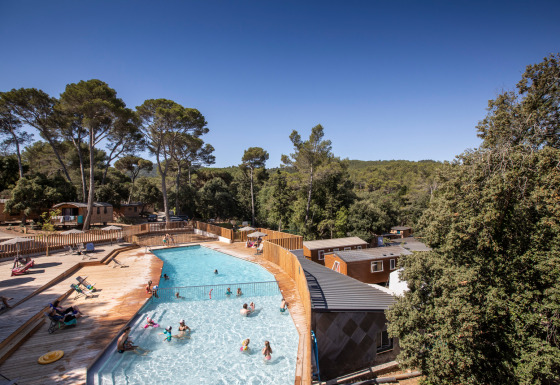 Outdoor swimming pool with wooden terrace and cabins at Huttopia La Forêt de Janas holiday park, surrounded by trees.