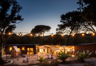 Aftenbillede af Huttopia La Forêt de Janas feriepark i Provence-Alpes-Côte d’Azur med lyskæder og legeplads.