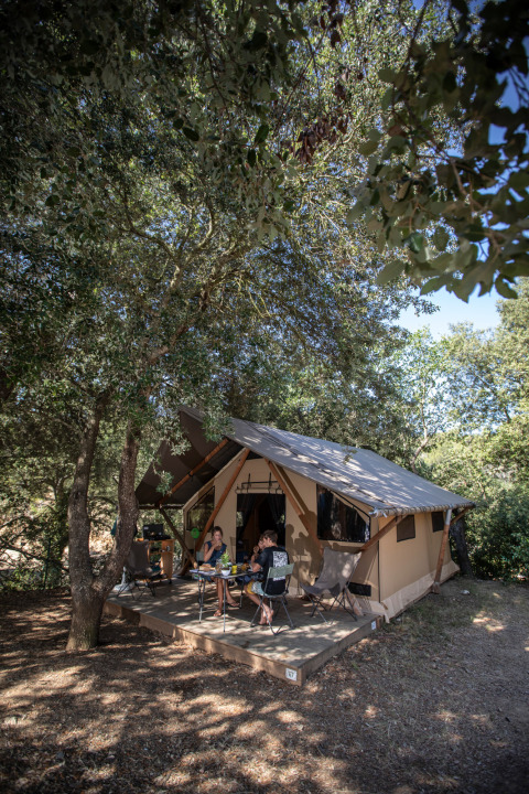 Familia relajándose frente a una tienda tipo safari entre árboles en Huttopia La Forêt de Janas, Francia.