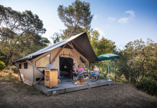 Gezin relaxt aan safaritent op houten terras met hond, parasol en bosomgeving in Zuid-Frankrijk.