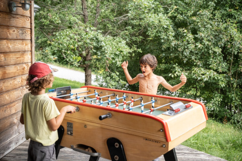 Dos niños juegan futbolín al aire libre junto a una cabaña de madera en Village Huttopia Pays de Condrieu, Francia.