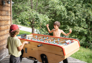 Zwei Kinder spielen Tischfußball im Freien an einer Holzhütte im Village Huttopia Pays de Condrieu, Frankreich.