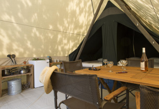 Interieur van een safaritent met tafel, stoelen, keukenblok, koelkast en een fles wijn op tafel.