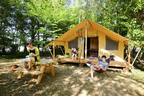 Tres personas relajándose y leyendo frente a una tienda safari Trappeur en un campamento arbolado en verano.