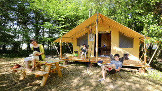 Tres personas relajándose y leyendo frente a una tienda safari Trappeur en un campamento arbolado en verano.
