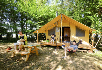 Tre personer slapper af og læser foran et Trappeur safari telt i en skovfyldt campingplads om sommeren.