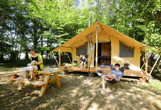 Tres personas relajándose y leyendo frente a una tienda safari Trappeur en un campamento arbolado en verano.
