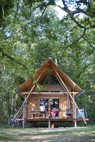 Tienda safari Cahutte en Village Huttopia Lac de Rillé, Francia, rodeada de árboles y personas descansando.