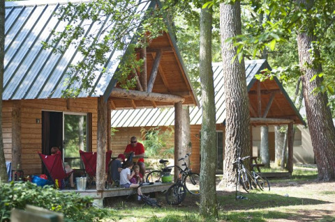 Familia descansa frente a una cabaña de madera en el bosque en Village Huttopia Lac de Rillé, Francia, cerca de bicicletas.