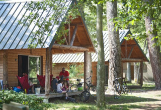 Family relaxes outside a wooden cabin in the forest at Village Huttopia Lac de Rillé, France, with bicycles nearby.