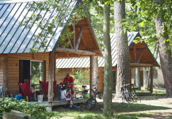 Family relaxes outside a wooden cabin in the forest at Village Huttopia Lac de Rillé, France, with bicycles nearby.