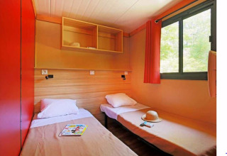 Zwei Einzelbetten, Fenster mit rotem Vorhang und offene Regale in Chalet Ottawa, Camping de Lyon, Frankreich.