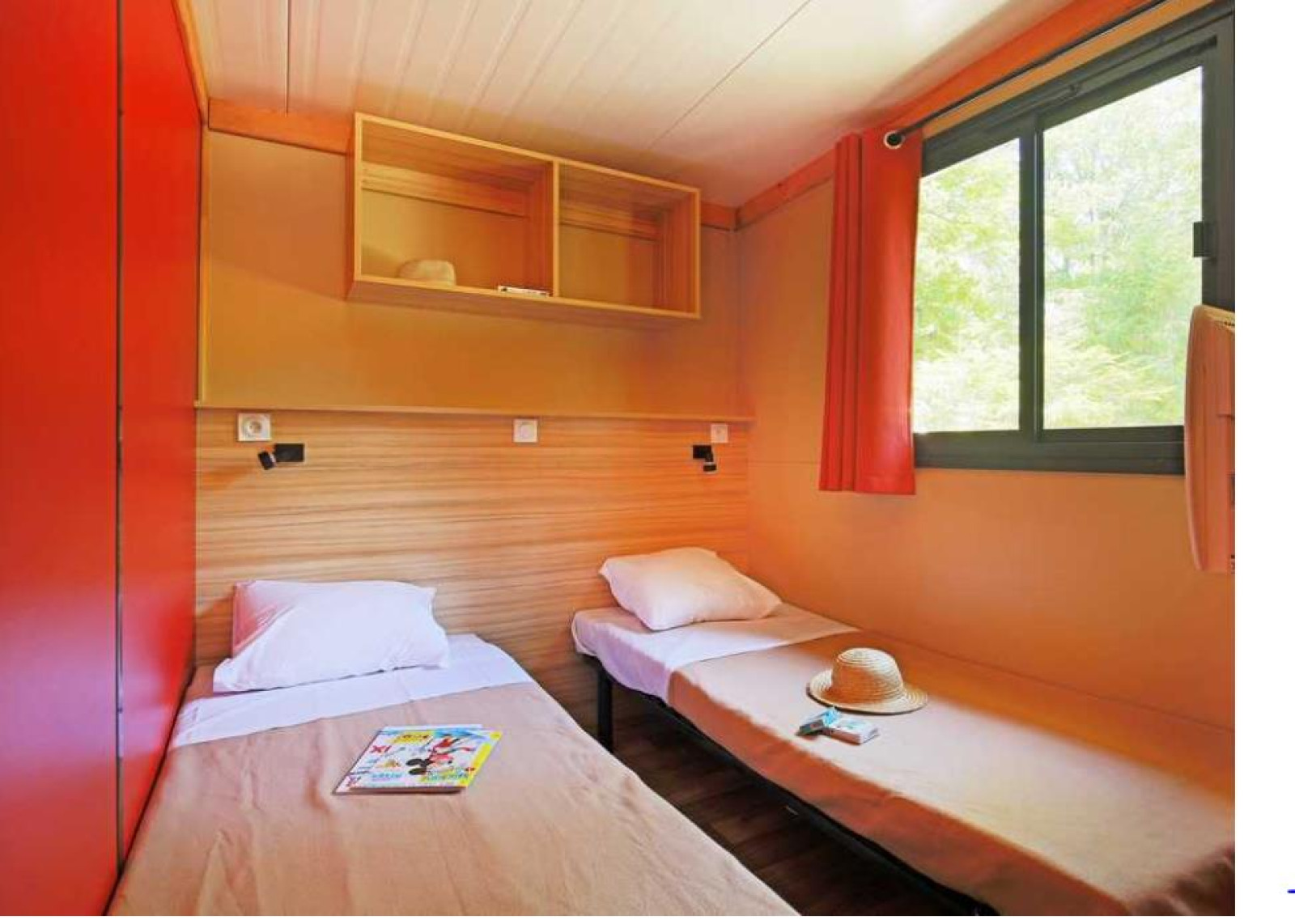 Twee eenpersoonsbedden, raam met rood gordijn en open planken in Chalet Ottawa bij Camping de Lyon, Frankrijk.