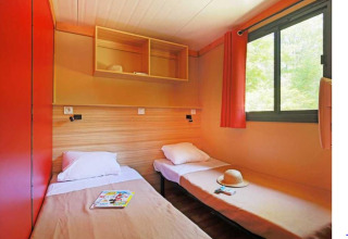 Dos camas individuales, ventana con cortina roja y estantes abiertos en Chalet Ottawa, Camping de Lyon, Francia.