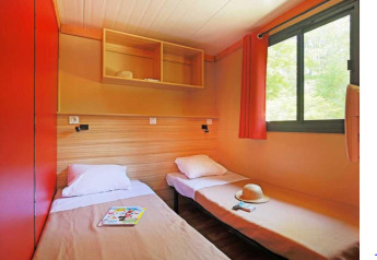 Due letti singoli, finestra con tenda rossa e scaffali aperti nello Chalet Ottawa al Camping de Lyon, Francia.