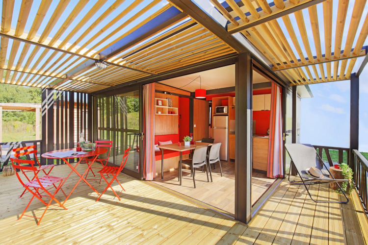 Luminoso interior de cabaña moderna con terraza de madera, muebles rojos y cocina-comedor abierta.