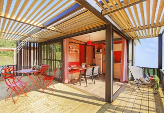Luminoso interior de cabaña moderna con terraza de madera, muebles rojos y cocina-comedor abierta.