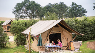 Una familia se sienta en una mesa frente a una tienda safari llamada Tent Trappeur, rodeada de naturaleza.