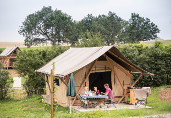 En familie sidder ved et bord foran en safari telt ved navn Tent Trappeur, omgivet af natur.