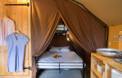 Interior de glamping con cama doble, litera, cortinas marrones, lavamanos, estantería y ropa colgada.