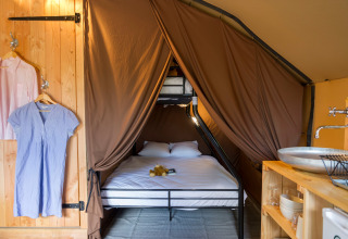 Gezellig glamping-interieur met dubbel bed, stapelbed, bruine gordijnen, lavabo en hangende kleren.