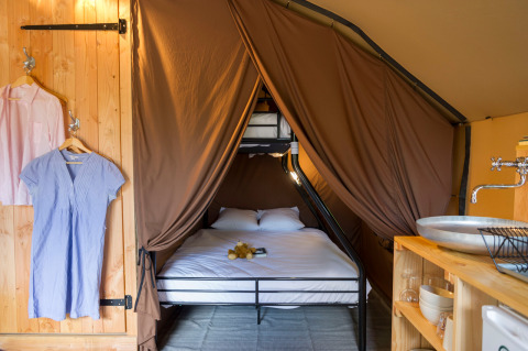 Cozy glamping tent interior with double bed, bunk bed, brown curtains, sink, wooden shelves, and clothes.