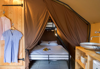 Gezellig glamping-interieur met dubbel bed, stapelbed, bruine gordijnen, lavabo en hangende kleren.