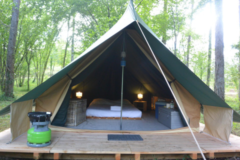 Interior of a comfortable teepee tent with bed, lamps, and storage set in a lush green forest.