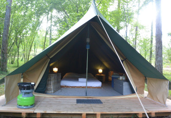 Interior of a comfortable teepee tent with bed, lamps, and storage set in a lush green forest.