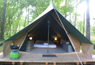 Interieur van een comfortabele tipi tent met bed, lampen en opslag in een groene bosrijke omgeving.