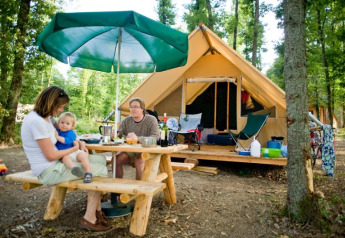 Family relaxes at a picnic table by a Canadienne safari tent at Village Huttopia Senonches, France.