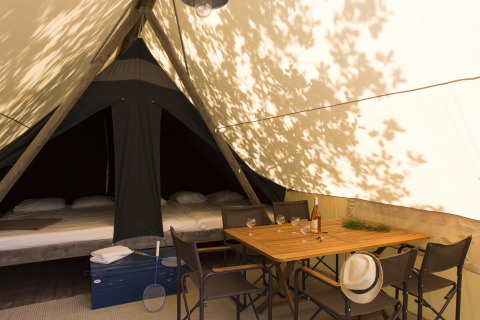 Binnenkant van een safaritent met bed, eettafel, stoelen, fles wijn en een hoed in de schaduw.