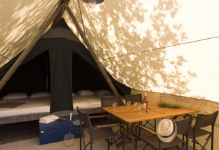 Interior de una tienda safari con cama, mesa de comedor, sillas, botella de vino y sombrero.